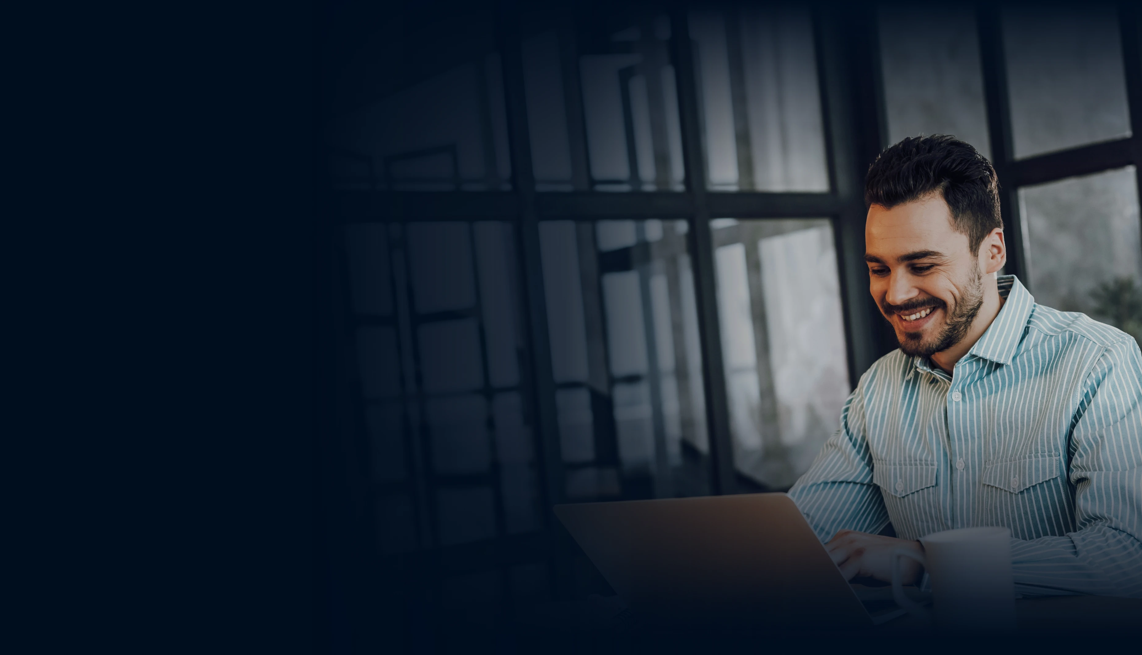 Smiling man in a striped shirt working on a laptop in a modern office setting.