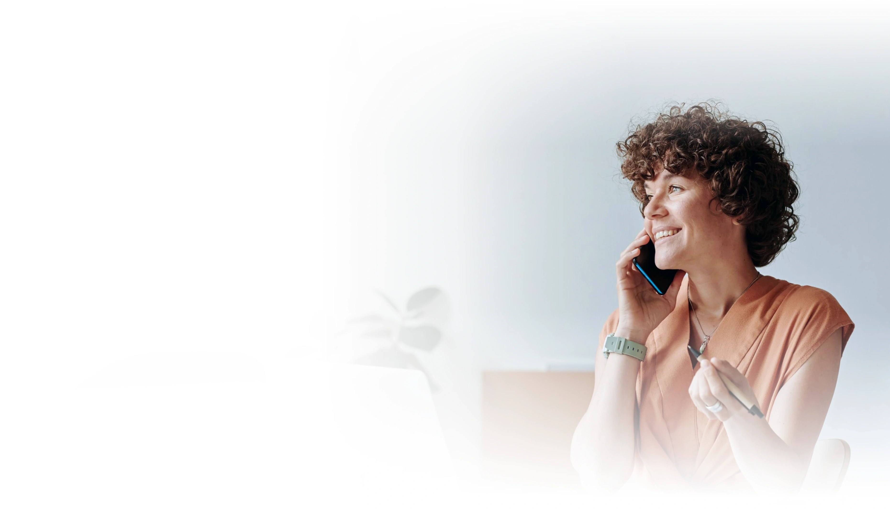 Smiling woman with curly hair talking on a smartphone in a bright, airy office setting.